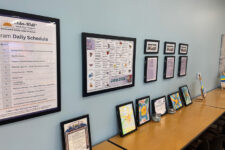 Wall-mounted schedule posters and framed artwork along a light blue wall, with a long wooden counter below exhibiting art frames and certificates.