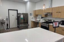 Break room with white island, stainless fridge, sink and stove along the wall, and a sign reads 'Employees Beyond This Point'.