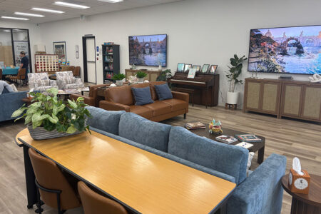 Open showroom/lounge with a blue sectional sofa and a brown leather sofa around wooden coffee tables; two large wall-mounted TVs show a river scene, with potted plants, a piano, and additional seating in the background.