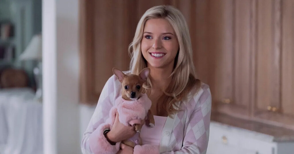 Smiling woman, Lexi Minetree, in a pink cardigan holding a small brown dog wearing a pink coat indoors in a bright kitchen setting.