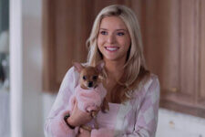 Smiling woman, Lexi Minetree, in a pink cardigan holding a small brown dog wearing a pink coat indoors in a bright kitchen setting.