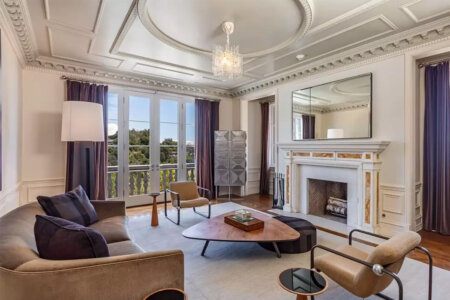 Elegant living room with chandelier, marble fireplace, large mirror, and purple-draped windows overlooking greenery outside balcony doors.