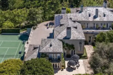 Bird's-eye view of a large gray-roofed mansion with a fenced green tennis court to the left and lush trees surrounding the property, all under a sunny sky.
