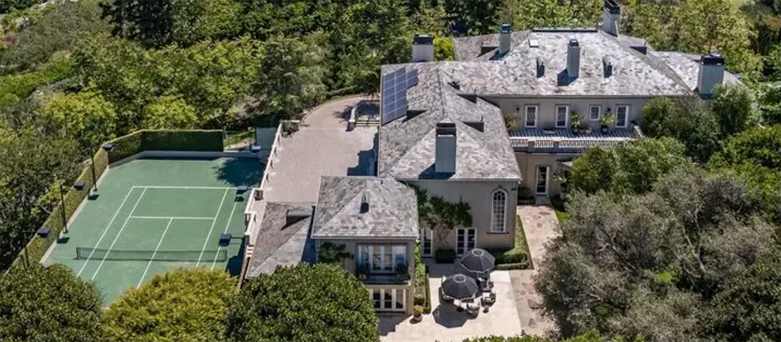 Bird's-eye view of a large gray-roofed mansion with a fenced green tennis court to the left and lush trees surrounding the property, all under a sunny sky.