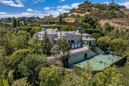 A luxury hillside home with a tennis court in a fenced yard, surrounded by trees under a bright blue sky with fluffy clouds.