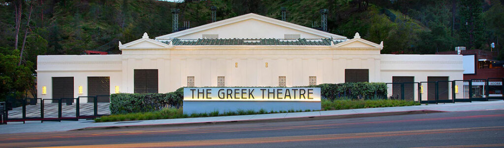 Front view of The Greek Theatre building, a white neoclassical venue with a sign reading THE GREEK THEATRE in front of a hedge.