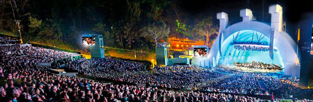 Outdoor night concert in an open-air amphitheater with a large audience and a blue-lit stage performing a symphony/chorus.