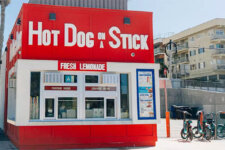 Red Hot Dog on a Stick stand with a large white sign and 'Fresh Lemonade' menu, set in a sunny urban lot with parked cars nearby.