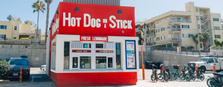 Red Hot Dog on a Stick stand with a large white sign and 'Fresh Lemonade' menu, set in a sunny urban lot with parked cars nearby.