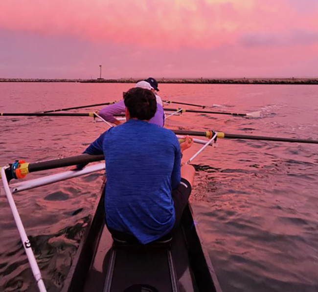 Two rowers in a racing boat on pink sunset water, viewed from behind