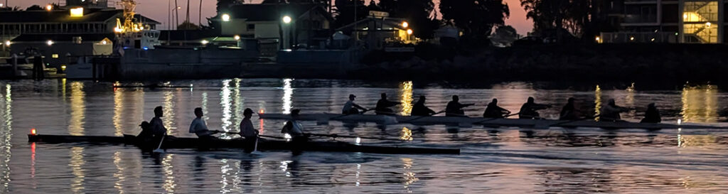 Rowing team in two long boats racing at dusk on a calm river with city lights reflecting on the water