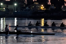 Rowing team in two long boats racing at dusk on a calm river with city lights reflecting on the water