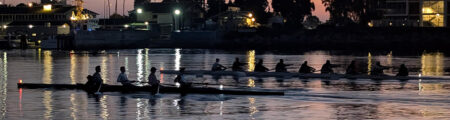 Rowing team in two long boats racing at dusk on a calm river with city lights reflecting on the water