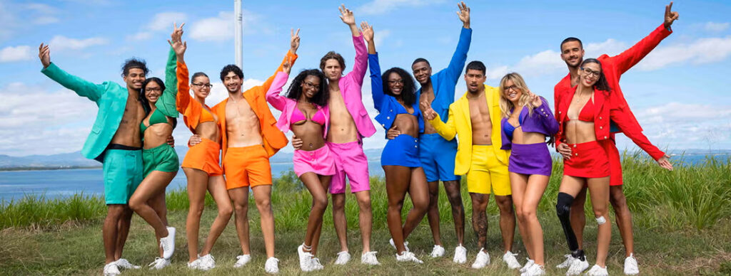 Group of diverse friends in neon-colored outfits posing outdoors with arms raised, against a blue sky and water.