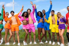 Group of diverse friends in neon-colored outfits posing outdoors with arms raised, against a blue sky and water.