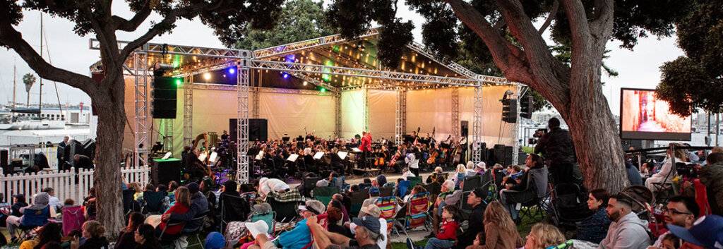 Outdoor concert on a lawn: a full stage with an orchestra performing under colorful lights, audience seated on blankets and chairs near trees by the water.