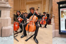 Young cellist standing outside Carnegie Hall with his cello next to a stone column and plaque (street scene).
