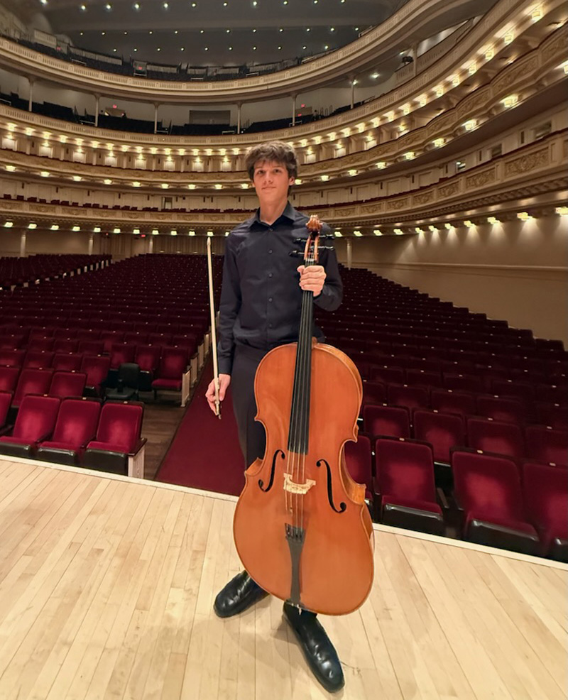 Cellist standing on stage in an empty concert hall, holding a cello and bow with red seats in the background.Cello on the floor in front of him and orchestral hall ambiance visible behind him.