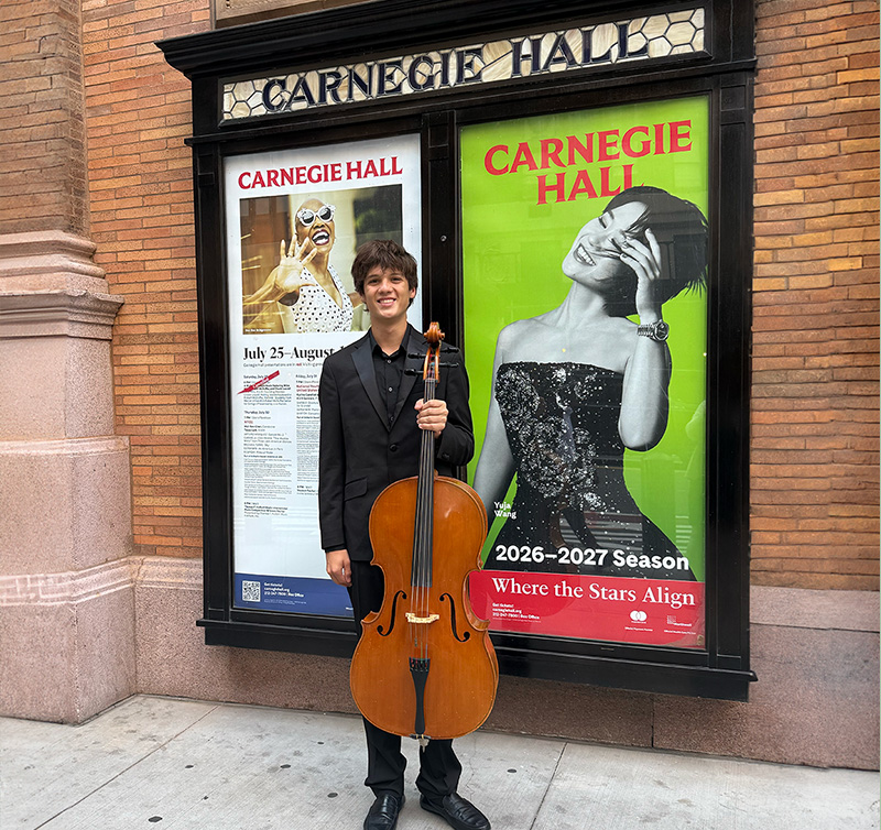 Young man in a black suit stands on a sidewalk, holding a cello in front of Carnegie Hall posters outside the venue.