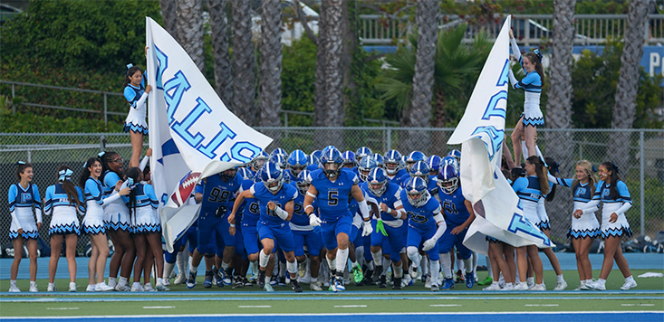 Blue football team bursts through large white banners at a stadium, flanked by cheerleaders in blue-and-white outfits on both sides.