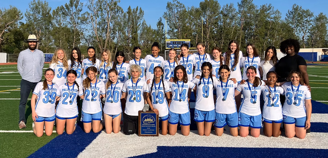 Girls soccer team posing on a football field with coaches, holding a plaque trophy after a championship win.