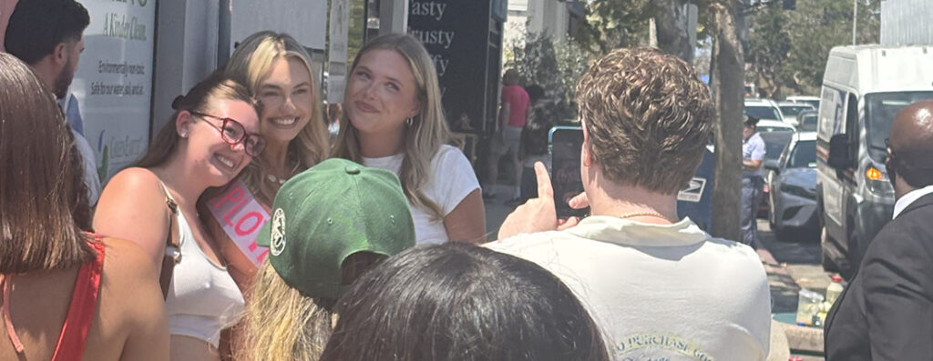 Group of young women smiling and posing for a photo on a sunny city sidewalk while a man takes their picture with a phone behind them.