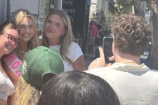 Group of young women smiling and posing for a photo on a sunny city sidewalk while a man takes their picture with a phone behind them.