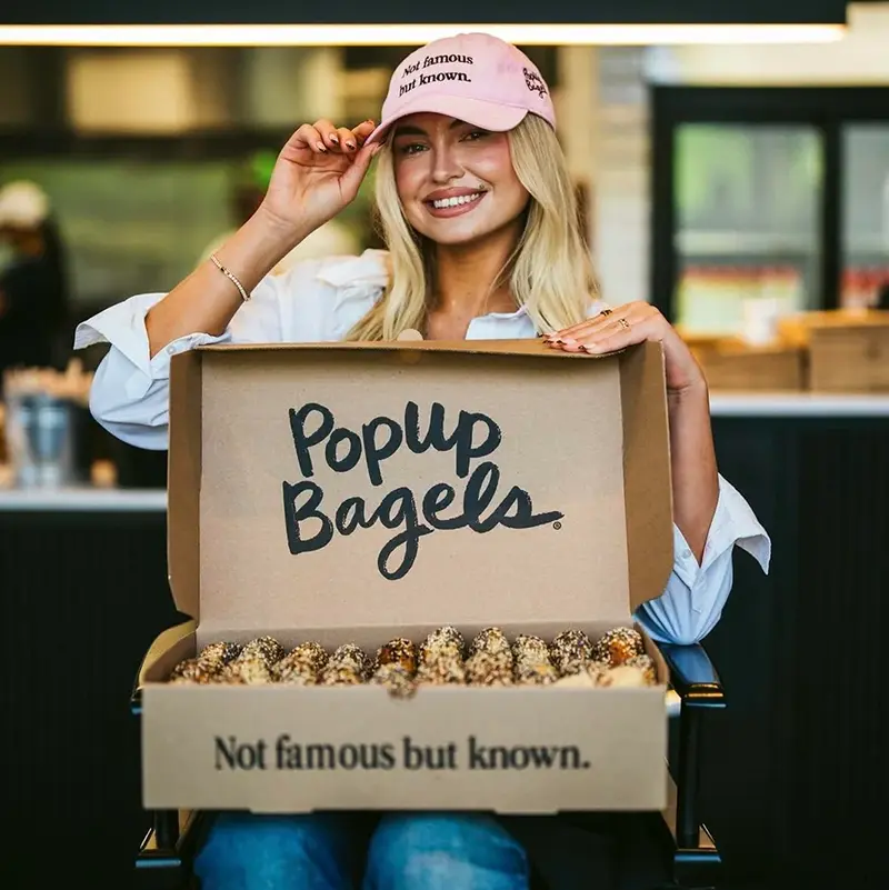 Woman smiling while holding an open box of PopUp Bagels in a cafe, wearing a pink hat that reads Not famous but known.