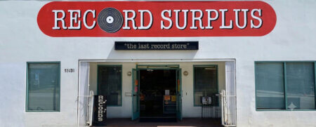 Storefront of a record shop with a red curved 'RECORD SURPLUS' sign, a black banner reading 'the last record store', and open teal doors.