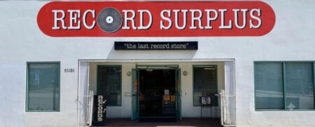 Storefront of a record shop with a red curved 'RECORD SURPLUS' sign, a black banner reading 'the last record store', and open teal doors.