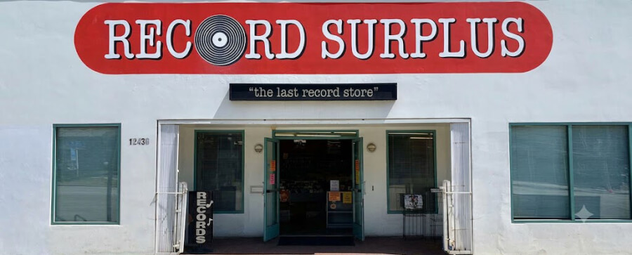Storefront of a record shop with a red curved 'RECORD SURPLUS' sign, a black banner reading 'the last record store', and open teal doors.