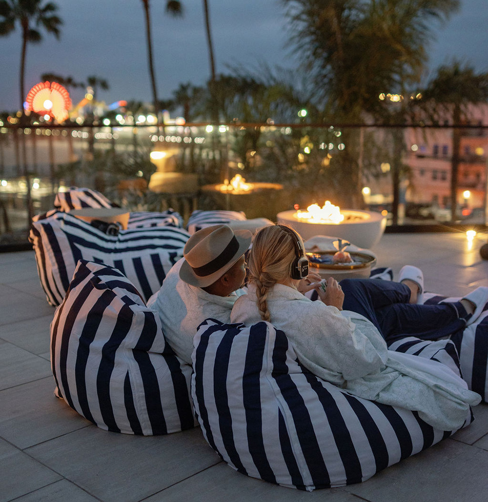 Couple lounging on striped outdoor cushions by a fire pit on a rooftop at night, wearing casual clothes and headphones, with city lights in the background.