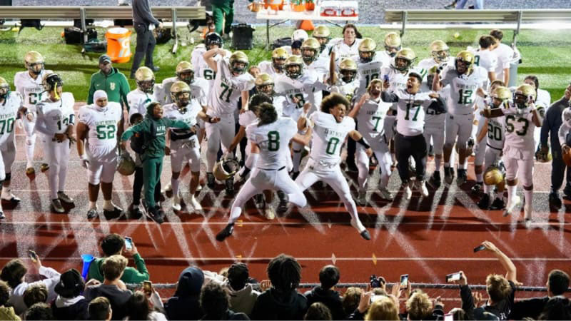 Football team in white uniforms leaps into the air in a group celebration on the track, as fans in the stands photograph the moment.