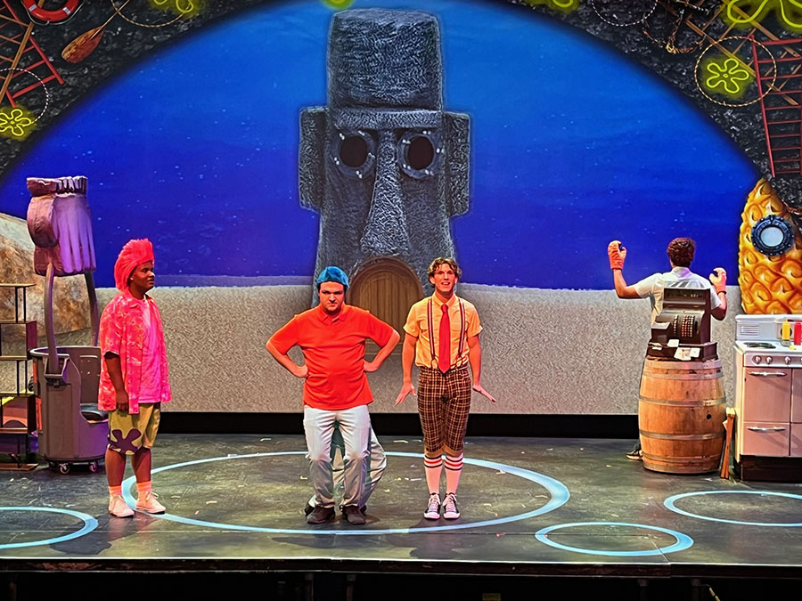 Three performers in bright costumes stand in a circular lit stage with a giant Moai backdrop and kitchen props on the right.