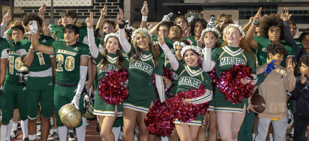 Group of football players and cheerleaders in green Mariners uniforms posing together on a track, holding red pom-poms and flashing peace signs.