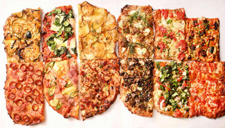 Assorted pizza slices in a row with various toppings like pepperoni, mushrooms, and greens on a light background.