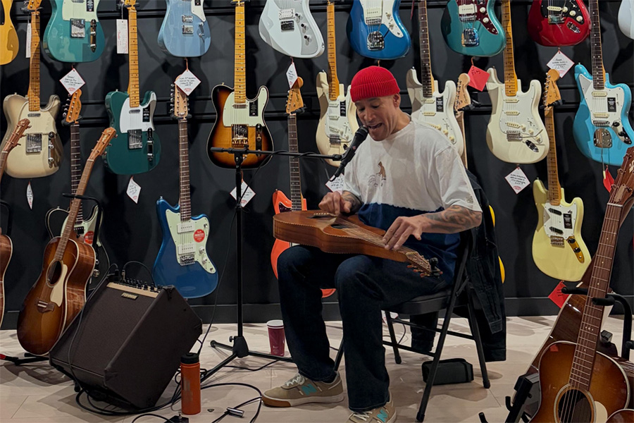 Musician in a red beanie playing a wooden string instrument on a chair, surrounded by colorful guitars hanging on the wall.