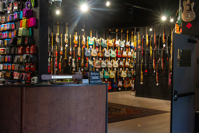 Interior of a music store with rows of colorful electric guitars hanging on the back wall and guitar accessories displayed on the left; a wooden counter is in the foreground.