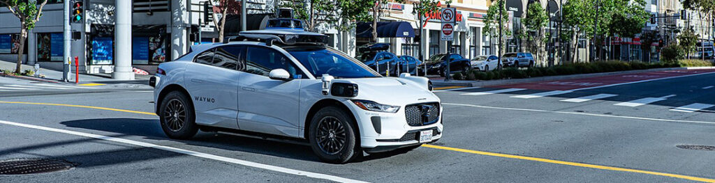 White Waymo self-driving car at an urban intersection with lidar sensors on the roof and 'WAYMO' branding on the side, waiting at a crosswalk.