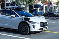 White Waymo self-driving car at an urban intersection with lidar sensors on the roof and 'WAYMO' branding on the side, waiting at a crosswalk.