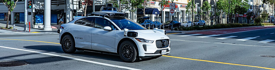 White Waymo self-driving car at an urban intersection with lidar sensors on the roof and 'WAYMO' branding on the side, waiting at a crosswalk.