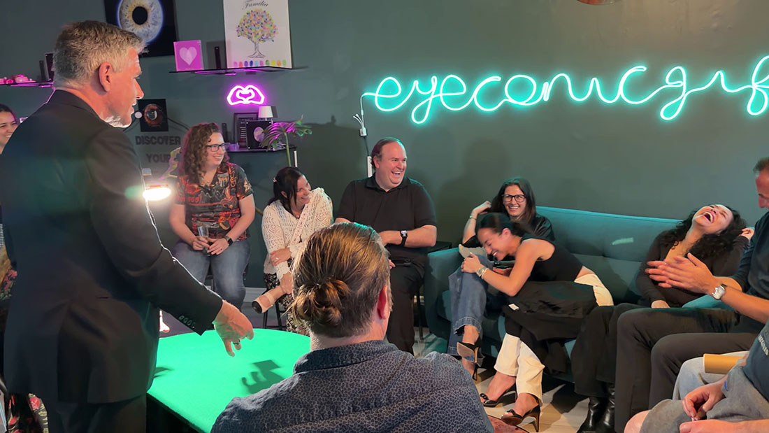 A group of adults sit on a teal couch and chairs, laughing as a man in a suit performs at a glowing green table in a room with neon signage on the wall.