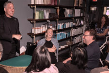 Man in a black blazer speaks to a small audience seated in a circle inside a store with shelves of hair products behind him.
