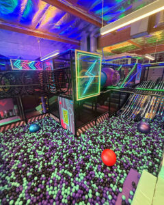 Colorful neon-lit indoor play area with a ball pit of purple and teal balls and modular climbing structures.9d1[1mNote:[0m no people visible in the scene.