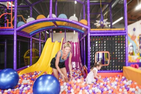 Woman and young girl playing in a colorful indoor ball pit with a purple climbing structure and yellow slide nearby.