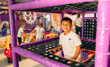 Smiling boy in a white shirt stands inside a purple indoor playground climbing frame, with other kids in the background.