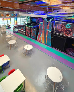 Colorful neon-lit arcade and seating area in a futuristic entertainment center with round white tables and clear chairs.