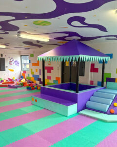 Indoor kids' play room with a purple canopy structure on a raised platform and teal steps, surrounded by colorful foam mats and blocks.