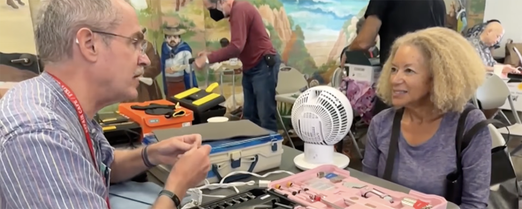 Man with glasses and a lanyard explains a tool kit to a smiling woman at a community workshop, with a pink tool case and a small fan on the table nearby.