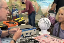 Man with glasses and a lanyard explains a tool kit to a smiling woman at a community workshop, with a pink tool case and a small fan on the table nearby.